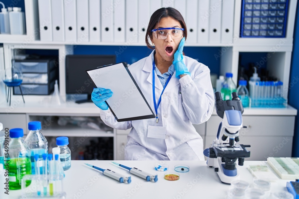 Hispanic young woman working at scientist laboratory afraid and shocked, surprise and amazed expression with hands on face