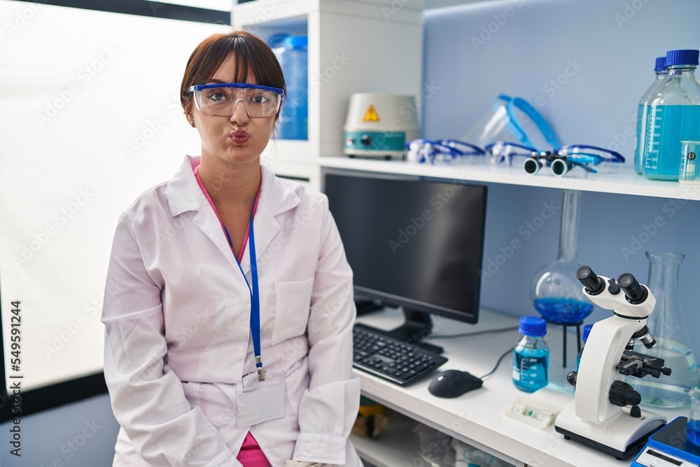 Young brunette woman working at scientist laboratory puffing cheeks with funny face. mouth inflated with air, crazy expression. Young brunette woman working at scientist laboratory puffing cheeks with funny face. mouth inflated with air, crazy expression.