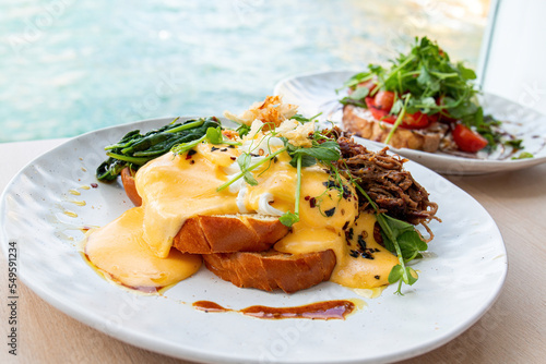 Healthy Breakfast Eggs Benedict with Whole meal Bread Toast, little salad and fresh herbs on wooden table in a café with view on beach at day sun light on background.