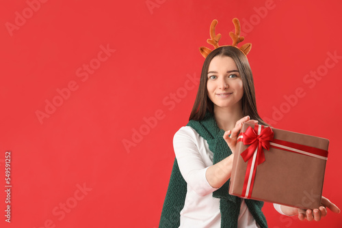 Young woman in reindeer horns with Christmas gift on red background