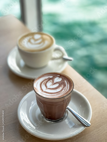 Close up Cup of Hot Chocolate with latte art on wooden table in a café with view on beach at day sun light on background.