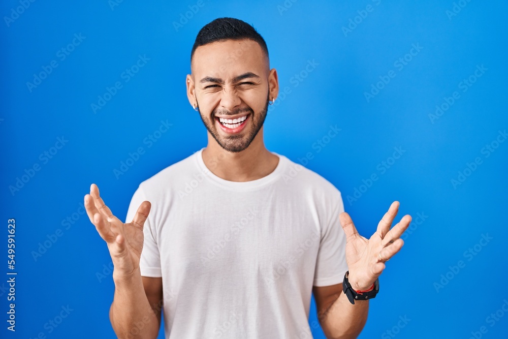 Fototapeta premium Young hispanic man standing over blue background celebrating mad and crazy for success with arms raised and closed eyes screaming excited. winner concept