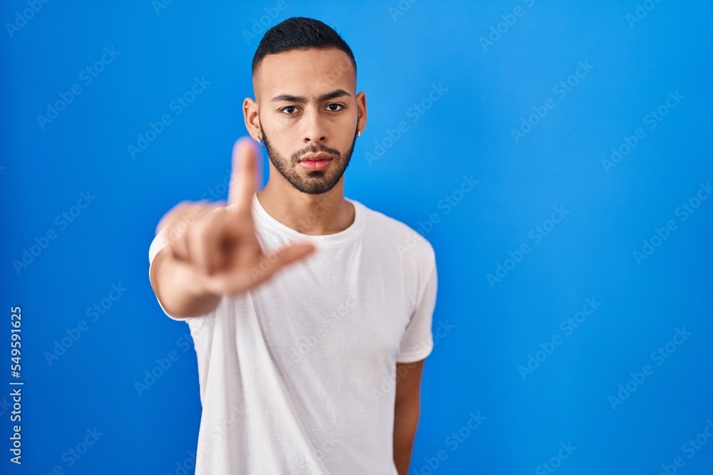Young hispanic man standing over blue background pointing with finger up and angry expression, showing no gesture Young hispanic man standing over blue background pointing with finger up and angry expression, showing no gesture