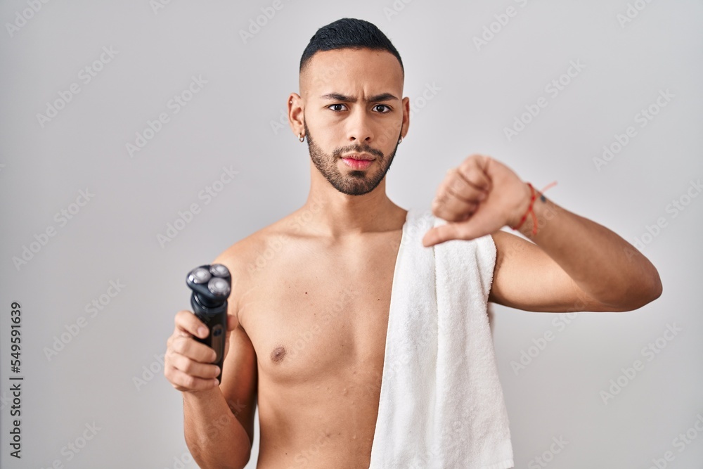 Fototapeta premium Young hispanic man holding electric razor with angry face, negative sign showing dislike with thumbs down, rejection concept
