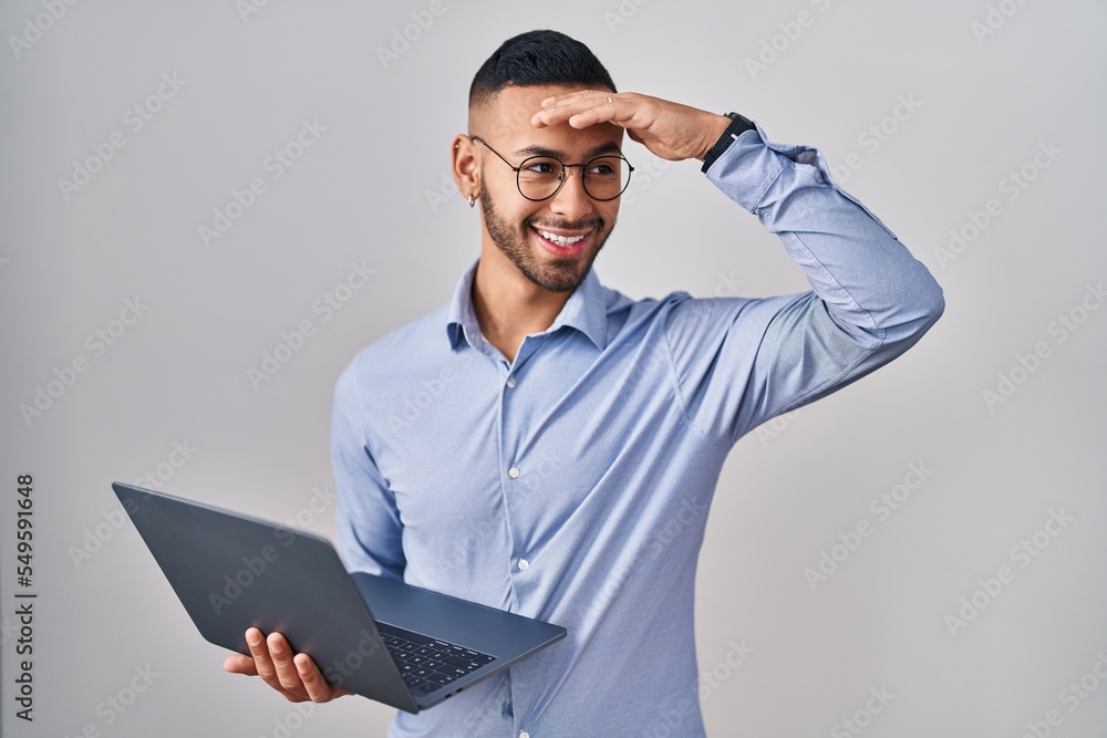 Young hispanic man working using computer laptop very happy and smiling looking far away with hand over head. searching concept. Young hispanic man working using computer laptop very happy and smiling looking far away with hand over head. searching concept.