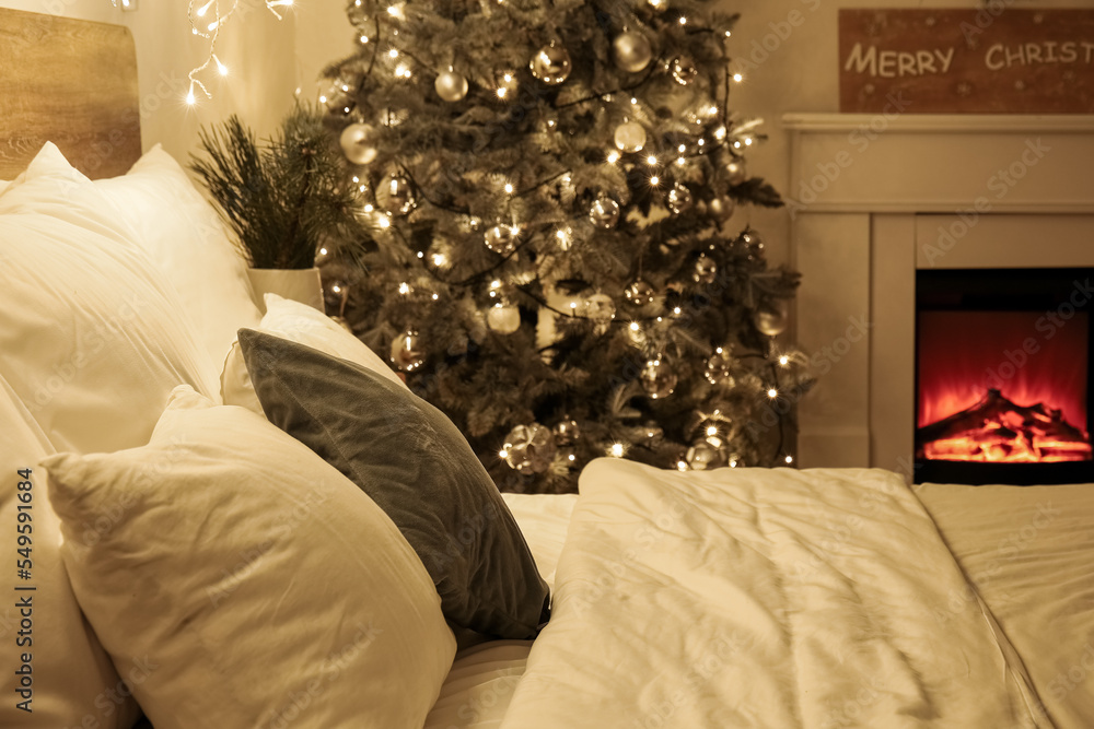 Fototapeta premium Big bed near glowing Christmas tree and fireplace in bedroom at night