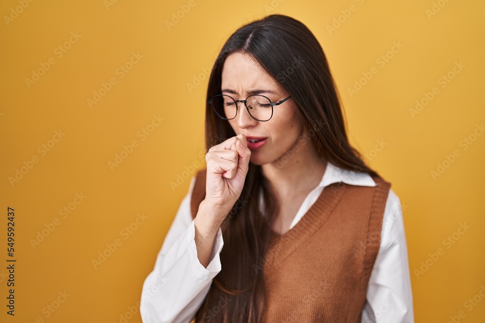 Young brunette woman standing over yellow background wearing glasses feeling unwell and coughing as symptom for cold or bronchitis. health care concept. Young brunette woman standing over yellow background wearing glasses feeling unwell and coughing as symptom for cold or bronchitis. health care concept.