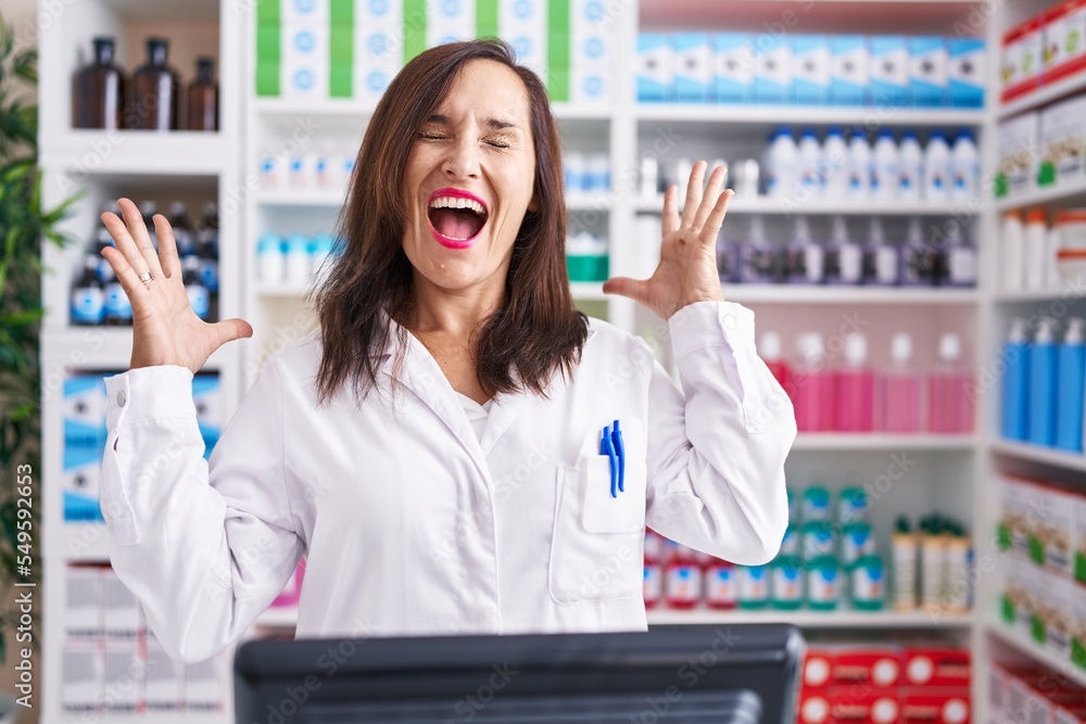 Middle age brunette woman working at pharmacy drugstore celebrating mad and crazy for success with arms raised and closed eyes screaming excited. winner concept