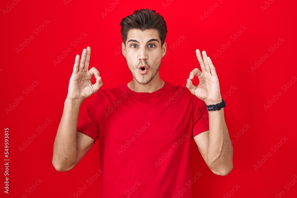Fototapeta premium Young hispanic man standing over red background looking surprised and shocked doing ok approval symbol with fingers. crazy expression