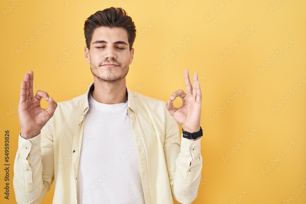 Young hispanic man standing over yellow background relaxed and smiling with eyes closed doing meditation gesture with fingers. yoga concept. Young hispanic man standing over yellow background relaxed and smiling with eyes closed doing meditation gesture with fingers. yoga concept.