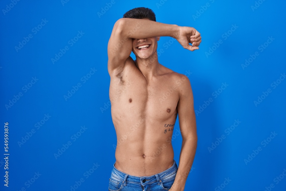 Young hispanic man standing shirtless over blue background covering eyes with arm smiling cheerful and funny. blind concept.