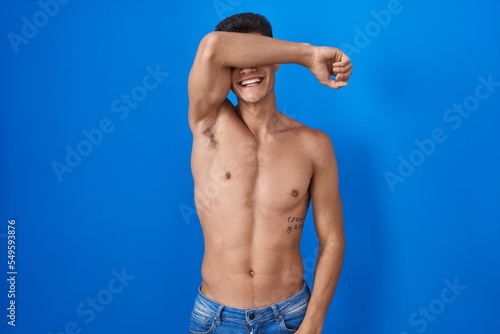 Young hispanic man standing shirtless over blue background covering eyes with arm smiling cheerful and funny. blind concept.