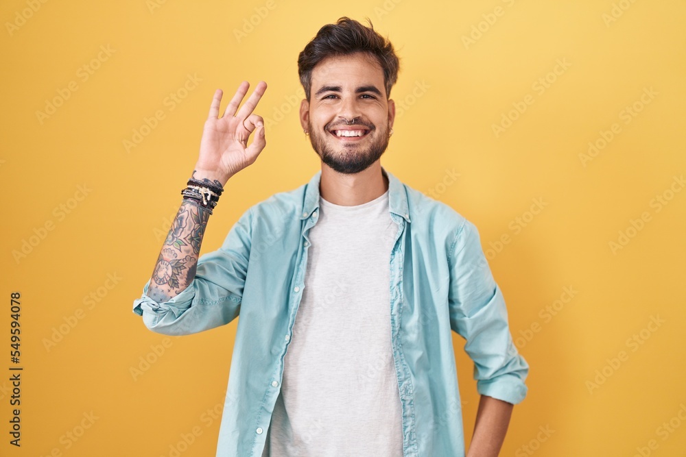Young hispanic man with tattoos standing over yellow background smiling positive doing ok sign with hand and fingers. successful expression. Young hispanic man with tattoos standing over yellow background smiling positive doing ok sign with hand and fingers. successful expression.