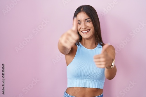 Young brunette woman standing over pink background approving doing positive gesture with hand, thumbs up smiling and happy for success. winner gesture. © Krakenimages.com