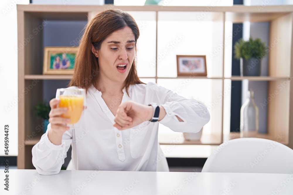 Brunette woman drinking glass of orange juice looking at the watch time worried, afraid of getting late Brunette woman drinking glass of orange juice looking at the watch time worried, afraid of getting late