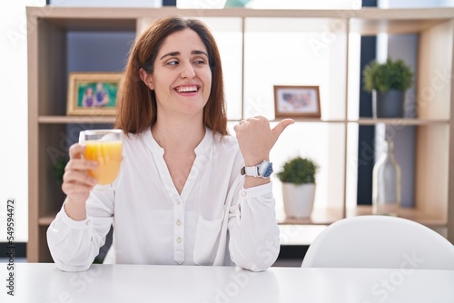 Brunette woman drinking glass of orange juice smiling with happy face looking and pointing to the side with thumb up.