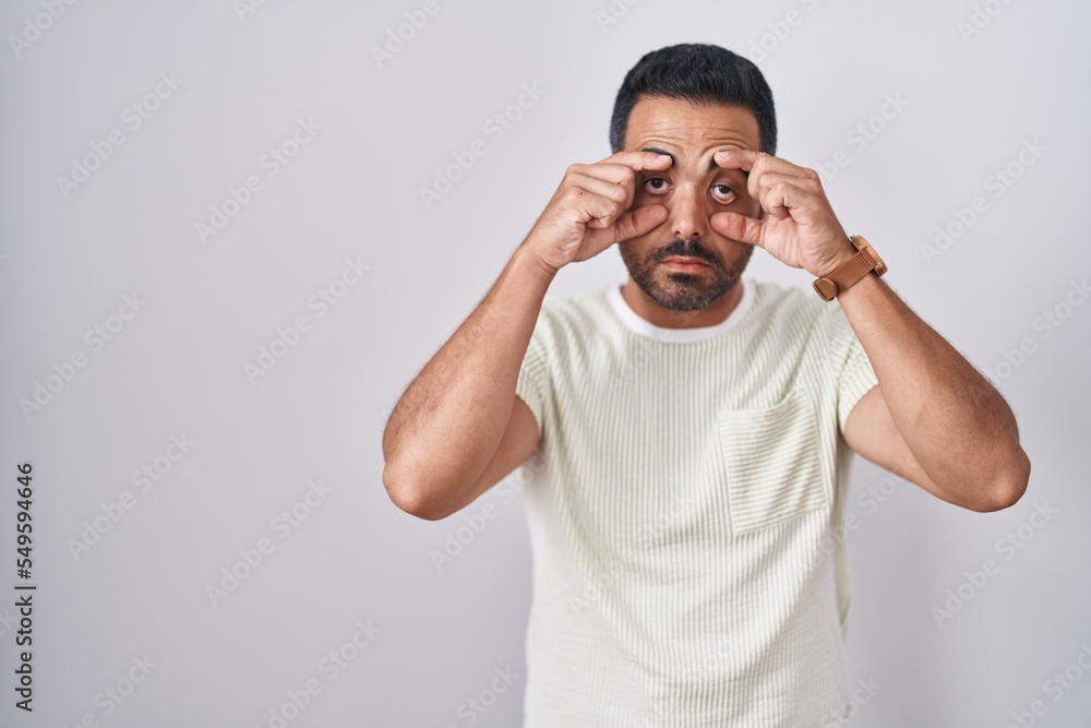 Hispanic man with beard standing over isolated background trying to open eyes with fingers, sleepy and tired for morning fatigue Hispanic man with beard standing over isolated background trying to open eyes with fingers, sleepy and tired for morning fatigue