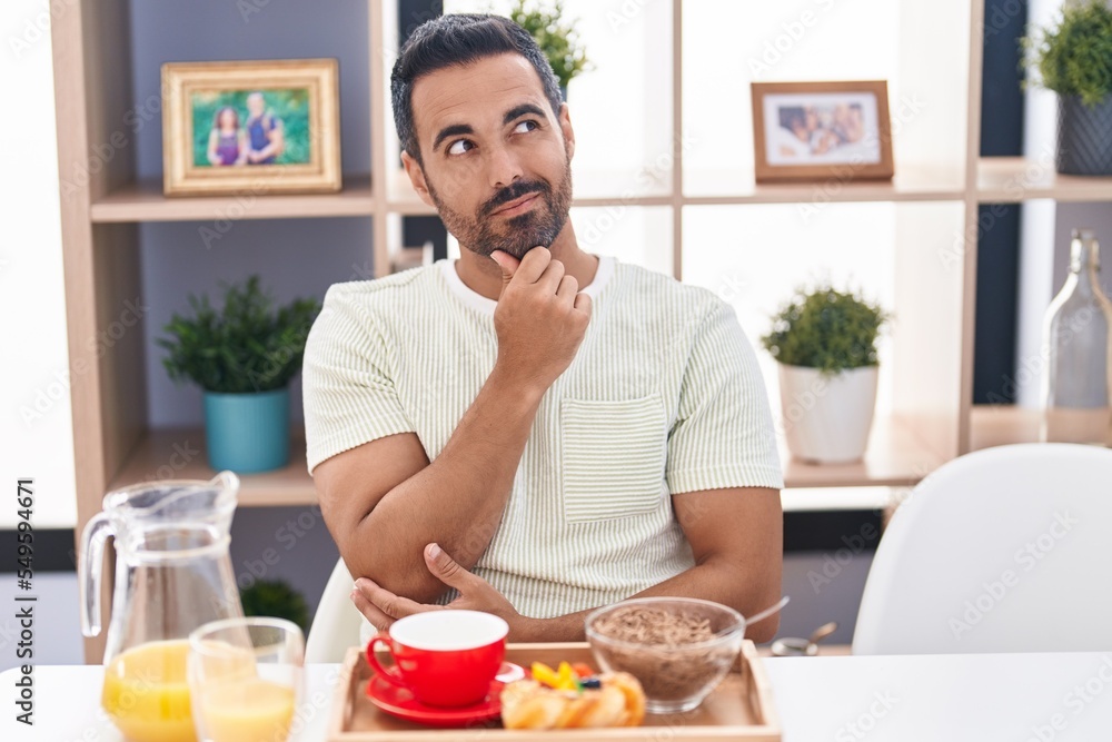 Hispanic man with beard eating breakfast thinking concentrated about doubt with finger on chin and looking up wondering Hispanic man with beard eating breakfast thinking concentrated about doubt with finger on chin and looking up wondering