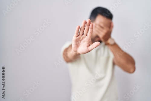 Hispanic man with beard standing over isolated background covering eyes with hands and doing stop gesture with sad and fear expression. embarrassed and negative concept.