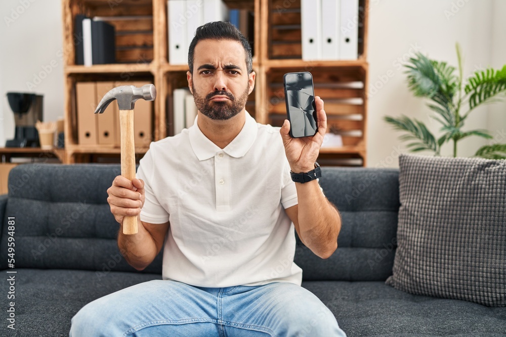 Young hispanic man with beard holding hammer and broken smartphone showing cracked screen depressed and worry for distress, crying angry and afraid. sad expression. Young hispanic man with beard holding hammer and broken smartphone showing cracked screen depressed and worry for distress, crying angry and afraid. sad expression.