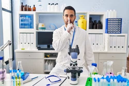 Young hispanic man with beard working at scientist laboratory asking to be quiet with finger on lips. silence and secret concept.