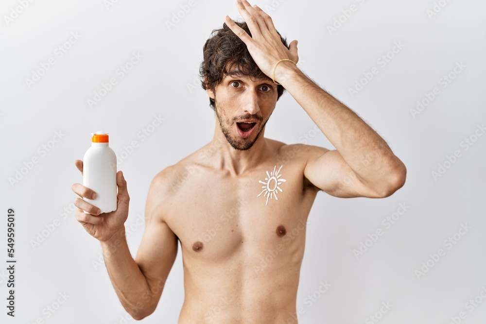 Fototapeta premium Young hispanic man standing shirtless holding sunscreen lotion surprised with hand on head for mistake, remember error. forgot, bad memory concept.