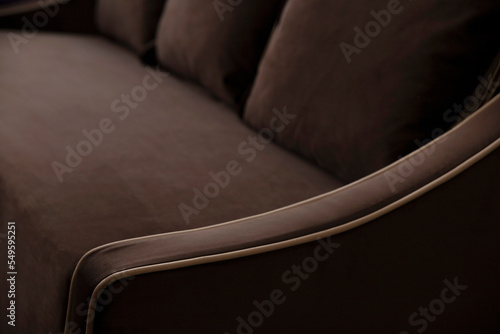 Defocused blurred background of a brown sofa with cushions and beige edging on the elbow of soft fleecy velor. Upholstered furniture close-up in deep shadows with copy space.