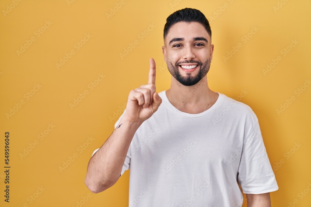 Young handsome man wearing casual t shirt over yellow background showing and pointing up with finger number one while smiling confident and happy. Young handsome man wearing casual t shirt over yellow background showing and pointing up with finger number one while smiling confident and happy.