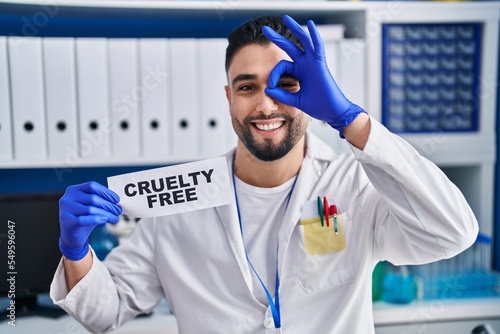 Young handsome man working at scientist laboratory holding cruelty free banner smiling happy doing ok sign with hand on eye looking through fingers