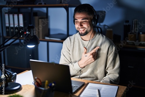 Young handsome man working using computer laptop at night cheerful with a smile of face pointing with hand and finger up to the side with happy and natural expression on face