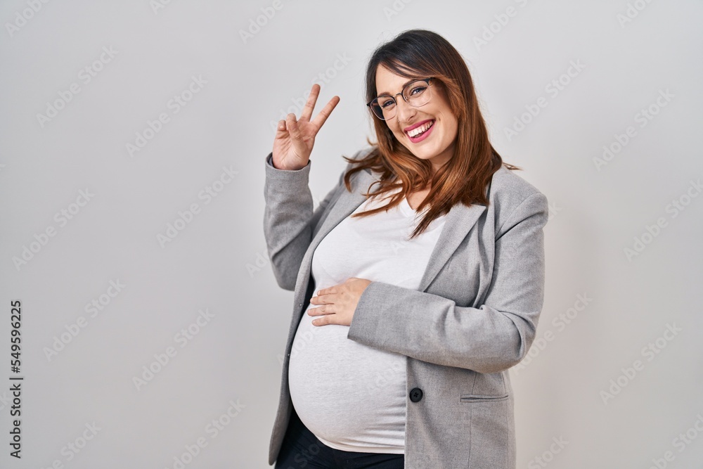 Pregnant business woman standing over white background smiling with happy face winking at the camera doing victory sign. number two. Pregnant business woman standing over white background smiling with happy face winking at the camera doing victory sign. number two.
