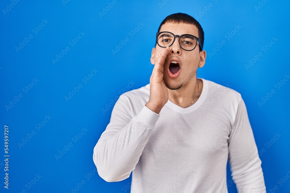 Young arab man wearing casual white shirt and glasses shouting and screaming loud to side with hand on mouth. communication concept. Young arab man wearing casual white shirt and glasses shouting and screaming loud to side with hand on mouth. communication concept.