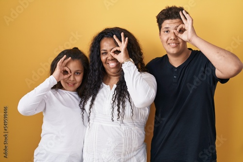 Family of mother, daughter and son standing over yellow background doing ok gesture with hand smiling, eye looking through fingers with happy face.