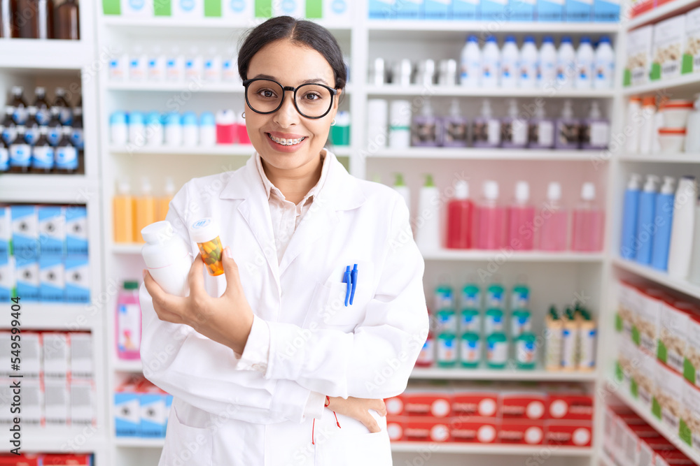 Young arab woman pharmacist holding pills bottles standing with arms crossed gesture at pharmacy