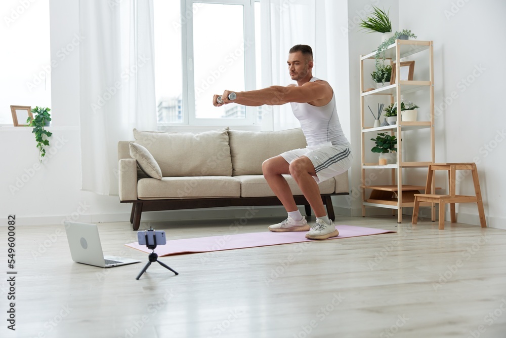 Man sports, watching a workout tape on his phone and repeating exercises sports blogger with dumbbells, pumped up man fitness trainer works out at home, the concept of health and body beauty Man sports, watching a workout tape on his phone and repeating exercises sports blogger with dumbbells, pumped up man fitness trainer works out at home, the concept of health and body beauty