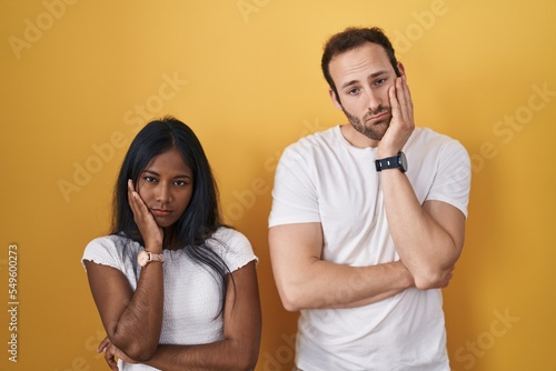 Interracial couple standing over yellow background thinking looking tired and bored with depression problems with crossed arms.