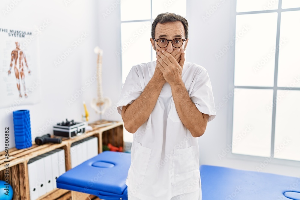 Middle age man with beard working at pain recovery clinic shocked covering mouth with hands for mistake. secret concept. Middle age man with beard working at pain recovery clinic shocked covering mouth with hands for mistake. secret concept.