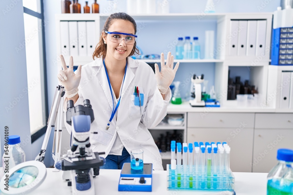 Young hispanic woman working at scientist laboratory showing and pointing up with fingers number eight while smiling confident and happy.