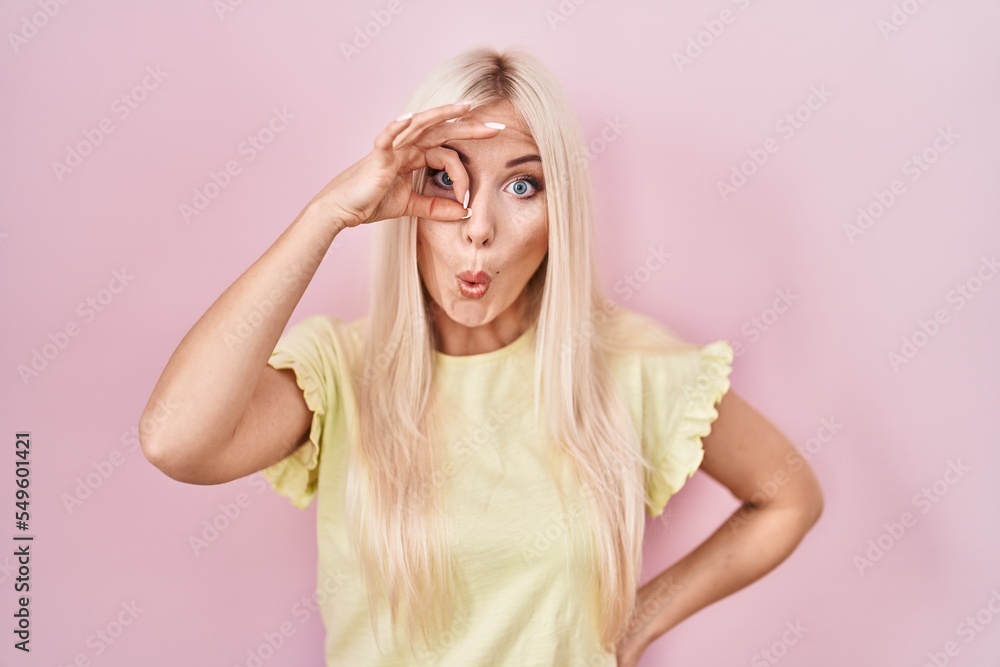 Caucasian woman standing over pink background doing ok gesture shocked with surprised face, eye looking through fingers. unbelieving expression.