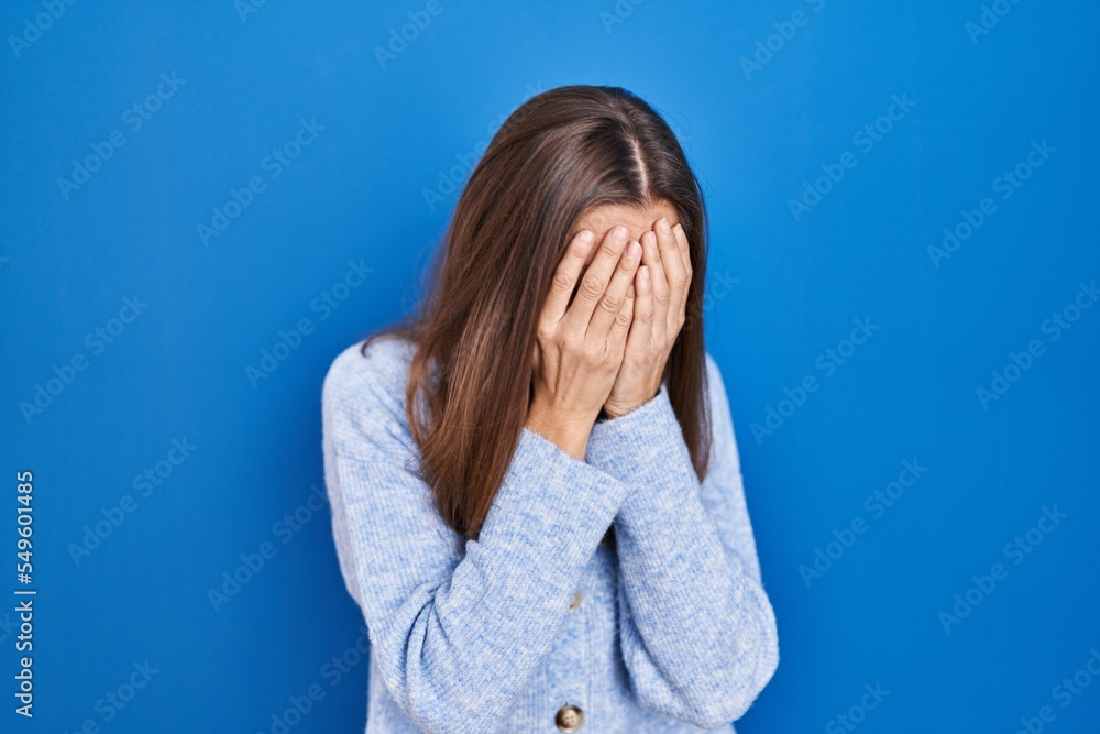 Young woman standing over blue background with sad expression covering face with hands while crying. depression concept. Young woman standing over blue background with sad expression covering face with hands while crying. depression concept.