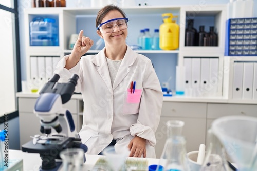 Hispanic girl with down syndrome working at scientist laboratory showing and pointing up with finger number one while smiling confident and happy.