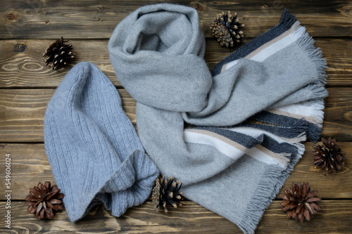 Striped grey cashmere scarf, tied in a knot. The knitted cap is gray. Pine cones. All objects lie on the surface of dark-colored wooden boards.