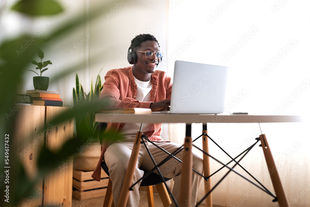 Fototapeta premium Long shot of young African American man wearing headphones working, studying at home using laptop. Copy space.