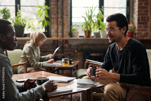 Focus on young businessman with cup of tea sitting in armchair by table in front of African American colleague and talking to him