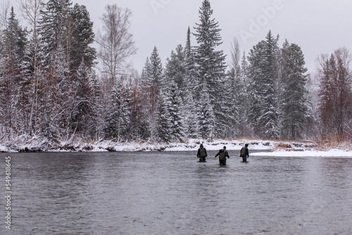 Group of fishermen on winter fishing. People in hiking gear are fording the river against the backdrop of a winter forest. Winter recreation and sports.