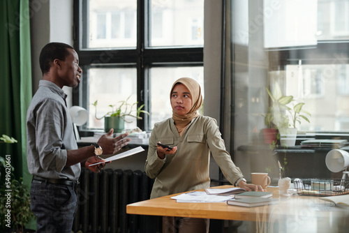 Young Muslim businesswoman listening to confident African American male colleague with document making report at meeting
