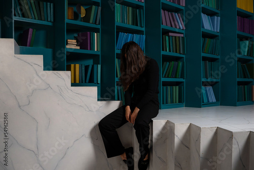 A sad young woman is sitting on a strange staircase against the background of bookshelves. Learning curve, concept. The first failures and loss of motivation.