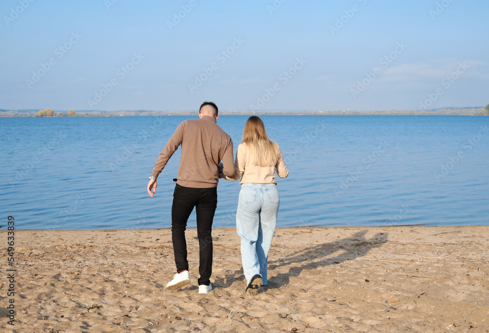 A young couple in jeans and sweaters hold hands and walk along the beach. Back view. Sunny weather A young couple in jeans and sweaters hold hands and walk along the beach. Back view. Sunny weather