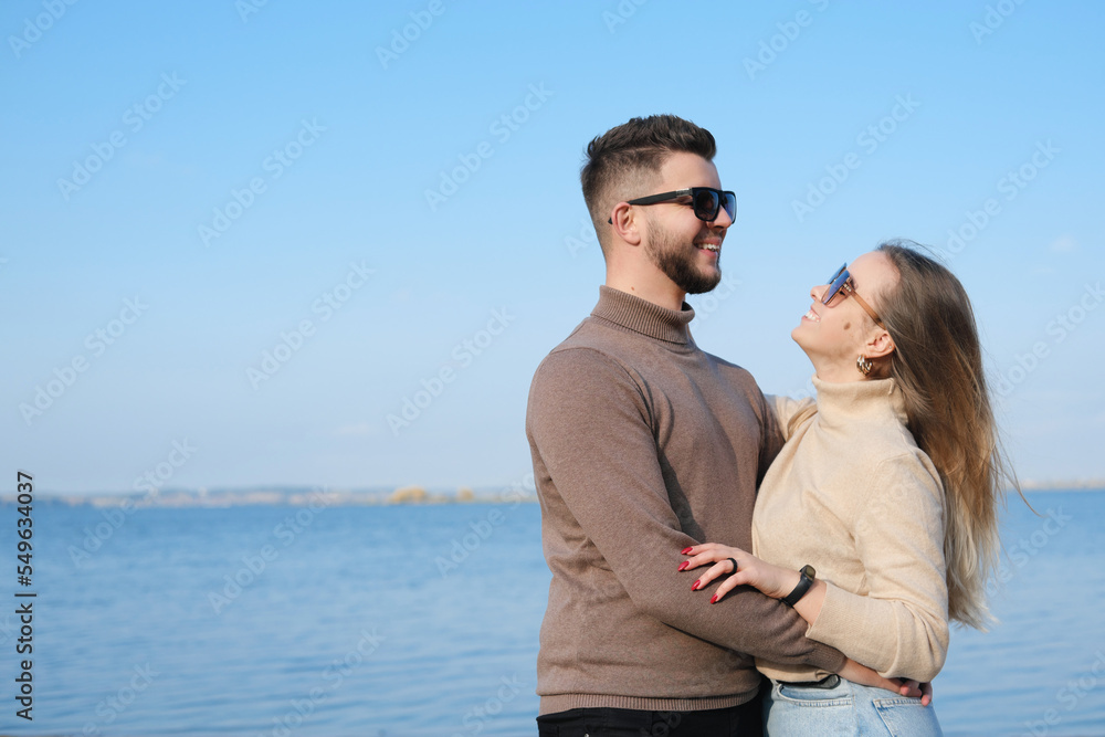 A young couple in jeans, sweaters and sunglasses stands on the beach near the lake and hugs A young couple in jeans, sweaters and sunglasses stands on the beach near the lake and hugs