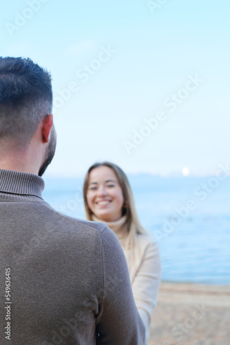 Young man and woman holding hands and posing on the beach against the backdrop of blue water. The girl looks at the guy and smiles. Focus on the guy's shoulder